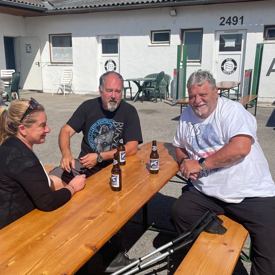 Drei Personen, zwei Männer und eine Frau, sitzen lächelnd um einen Holztisch und schauen in die Kamera. Auf dem Tisch stehen vier Bierflaschen. Hinter ihnen steht ein Gebäude mit einem Schild, das 2491 zeigt.