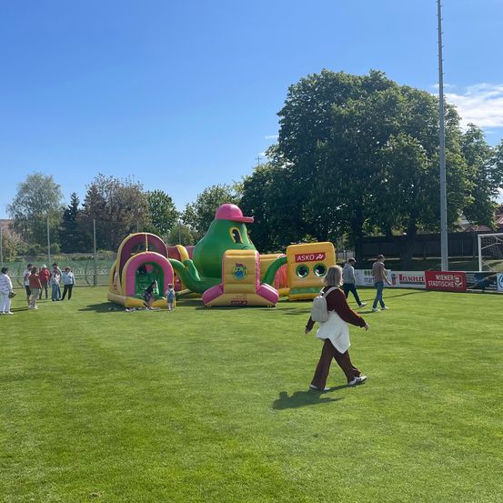 Ein großer aufblasbarer Spielplatz mit einer grünen und gelben schildkrötenartigen Struktur ist auf einem Rasenfeld aufgebaut. Mehrere Kinder spielen darin. Im Hintergrund stehen und gehen einige Personen herum. Bäume und ein Zaun sind in der Ferne sichtbar.