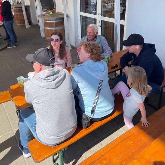 Eine Familie und Freunde sitzen zusammen an einem Tisch, lachen und genießen Getränke. Sie sind draußen in der Nähe eines Gebäudes.