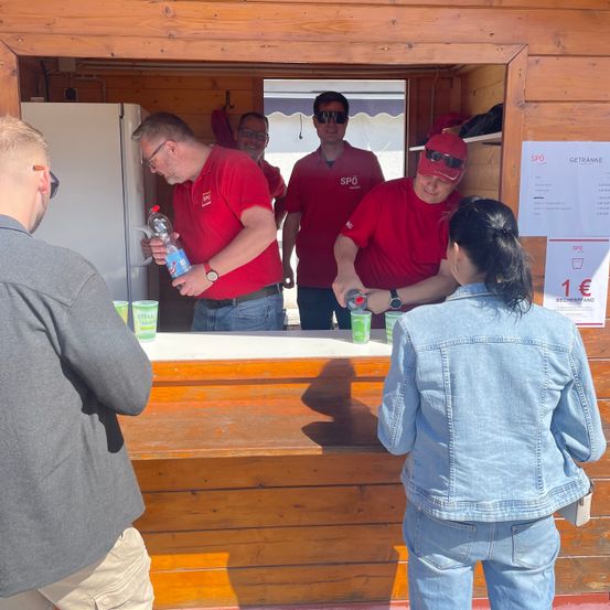 Eine Gruppe von Männern in roten Shirts bereitet Getränke an einem Tresen zu. Eine Frau in einer Jeansjacke wartet auf ihre Bestellung. Ein Kühlschrank und ein Preisschild sind sichtbar.