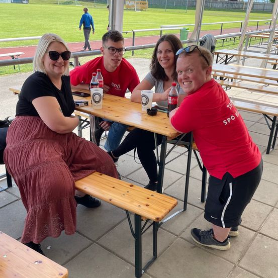 Vier Menschen sitzen lächelnd unter einem Zelt an einem Tisch. Eine Person lehnt sich auf den Tisch, während eine andere eine Coca-Cola-Flasche hält. Eine Person in einem roten Shirt steht in der Nähe.