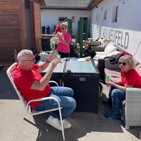 Zwei Menschen in roten Shirts sitzen draußen, einer mit einem Becher. Eine Frau in Pink hält hinter ihnen eine grüne Packung. Ein Tisch mit Glasplatte steht zwischen ihnen.