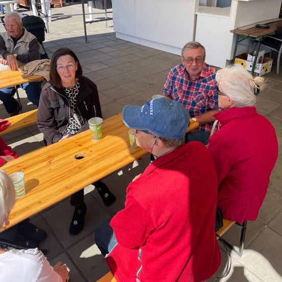 Eine Gruppe von Menschen sitzt auf Bänken in einem schattigen Außenbereich. Sie tragen Winterkleidung und einige haben eine Brille. Auf dem Tisch vor ihnen stehen Tassen.