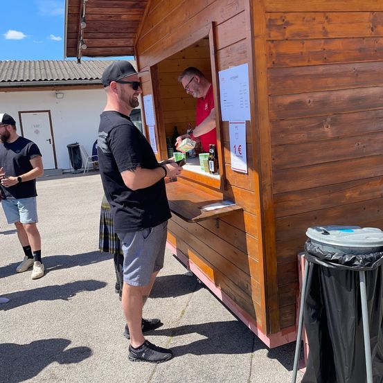 Ein Mann mit Hut steht vor einem Holzstand und kauft etwas von einem Mann in einem roten Shirt. Andere Leute stehen herum und ein Mülleimer ist in der Nähe.