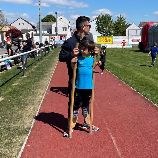 Ein Mann mit einer Prothese und ein Kind stehen auf einer Laufbahn. Zuschauer beobachten von der Seitenlinie, und ein Fußballfeld ist im Hintergrund.