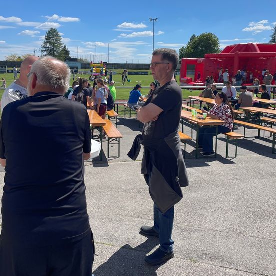 Eine Outdoor-Veranstaltung mit Tischen und Bänken, ein Mann mit Brille steht, eine aufblasbare Hindernisbahn im Hintergrund.