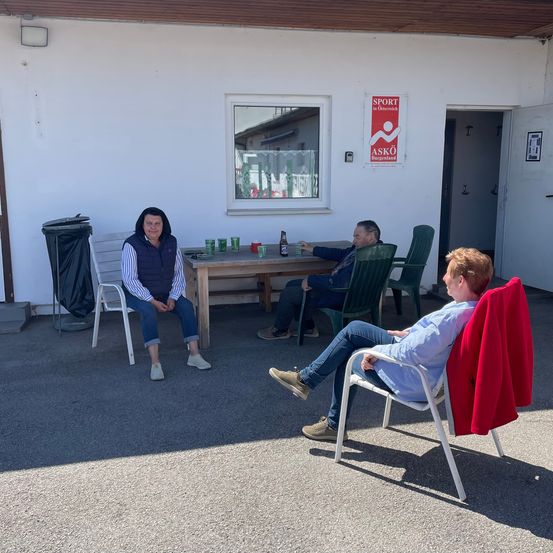 Vier Personen sitzen draußen, zwei Frauen und zwei Männer, im Gespräch. Ein Tisch hat Flaschen und Becher. Ein Schild mit der Aufschrift 'SPORT in Österreich' ist an der Wand angebracht.