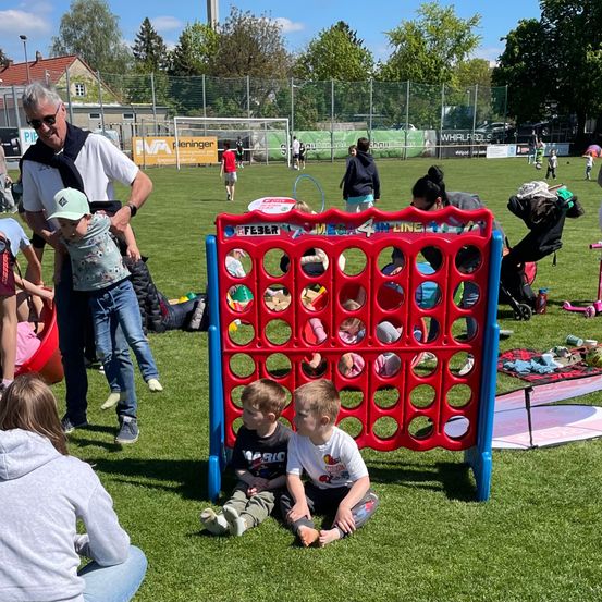 Eine Gruppe von Menschen spielt auf einem Feld. Ein Mann hält ein Kind, während zwei Jungen auf dem Boden sitzen. In der Nähe schiebt eine Frau einen Kinderwagen.