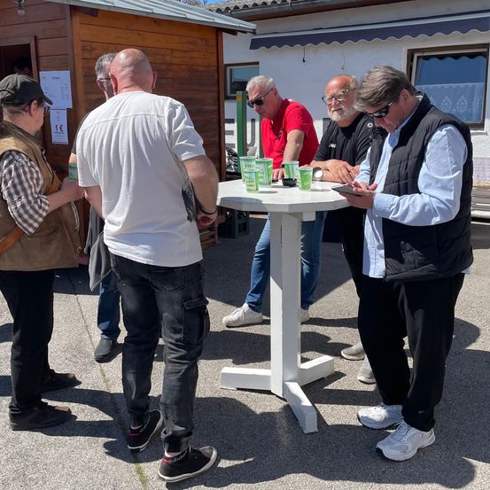 Eine Gruppe von Männern steht um einen weißen Tisch mit grünen Bechern, einer benutzt sein Handy. Sie sind lässig gekleidet. Dahinter steht ein hölzerner Stand.