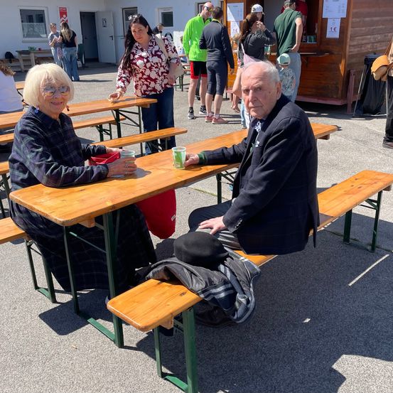 Zwei ältere Erwachsene sitzen auf einer Bank in einem Outdoor-Essbereich. Die Frau hält einen Becher, und der Mann trägt einen Hut. Dahinter versammeln sich Menschen in der Nähe eines Imbisswagens.