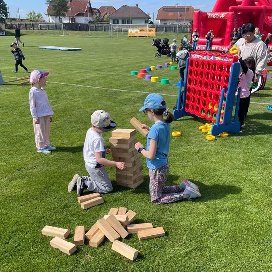 Kinder spielen auf einem Feld mit verschiedenen Spielzeugen, einschließlich eines großen Spiels von Connect Four und einem Stapel Holzblöcke. Eine Frau hilft einem Kind neben dem Spiel.
