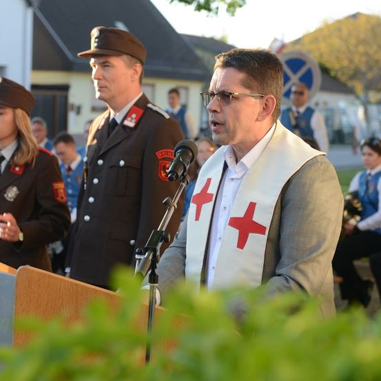 Ein Mann mit Brille spricht an einem Podium, trägt eine rote Kreuz-Schürze, mit einem uniformierten Beamten hinter ihm und Zuschauern in der Nähe.