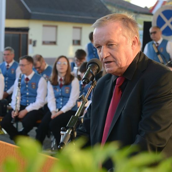 Ein Mann in Anzug und Krawatte spricht in ein Mikrofon bei einer Veranstaltung im Freien, mit einer Band in blauen Westen, die hinter ihm sitzen.