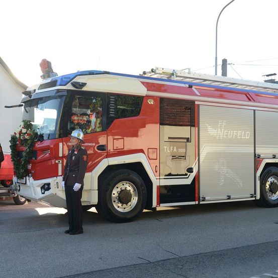 Ein Feuerwehrmann steht neben einem roten Feuerwehrauto mit dem Wort NEUFELD auf der Seite. Das Auto ist am Straßenrand geparkt.