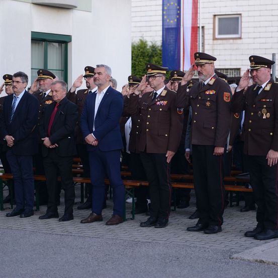 Männer in Militäruniformen und Anzügen stehen in einer Reihe und salutieren möglicherweise bei einer formellen Veranstaltung. Im Hintergrund befindet sich ein Gebäude mit EU- und nationalen Flaggen.