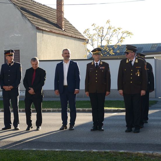 Fünf Männer in Uniformen stehen in einer Reihe vor einem Gebäude. Ein Mann im Anzug steht in der Mitte.