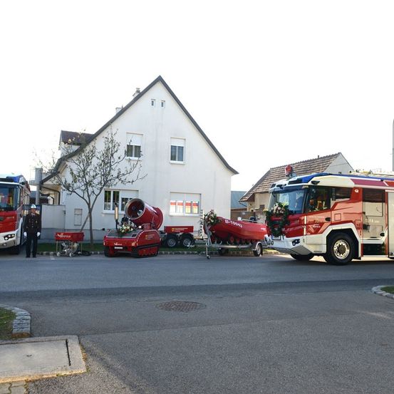 Mehrere rote Feuerwehrfahrzeuge sind in einer Reihe geparkt, eines mit einer roten Sirene auf dem Dach, ein anderes mit einem Bootsanhänger, vor einem weißen Haus mit Blumen auf der Vorderseite.
