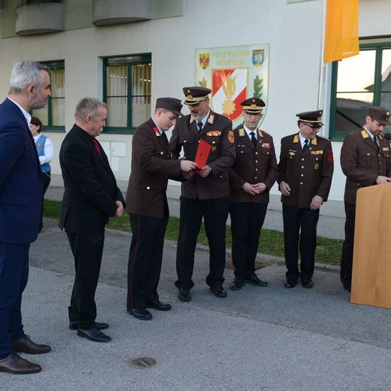 Eine Gruppe uniformierter Männer und ein Mann im Anzug tauschen Dokumente aus. Sie stehen vor einem Gebäude mit einem Banner und einer Flagge.