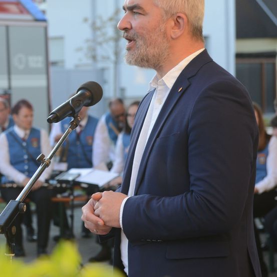 Ein Mann spricht in ein Mikrofon, formell gekleidet. Hinter ihm sitzen Musiker in blauen Westen. Im Hintergrund steht ein Baum und ein Gebäude.