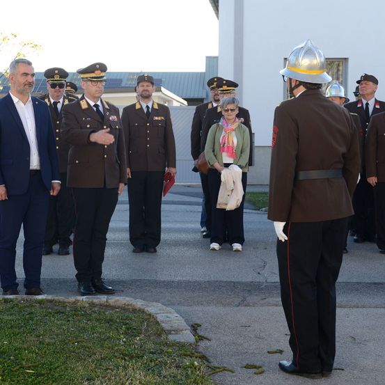 Mehrere Menschen stehen vor einem Gebäude. Ein Mann im Anzug und eine Frau mit einer Schürze sind unter ihnen. Einige Männer tragen Militäruniformen. Ein anderer Mann trägt einen Helm.