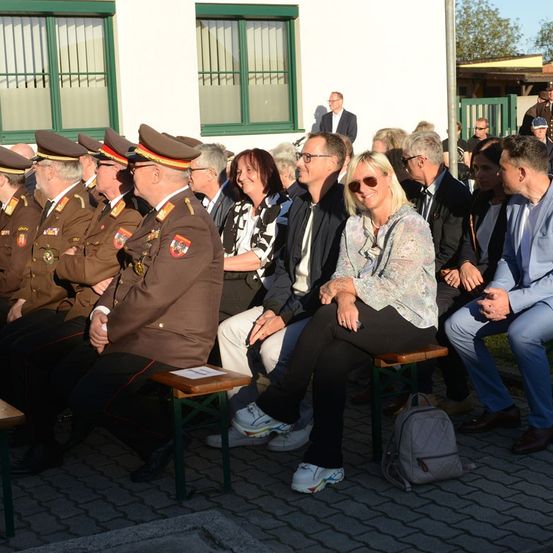 Eine Gruppe von Militärangehörigen und Zivilisten sitzt zusammen in hölzernen Stühlen vor einem Gebäude. Sie scheinen an einer Zeremonie teilzunehmen.