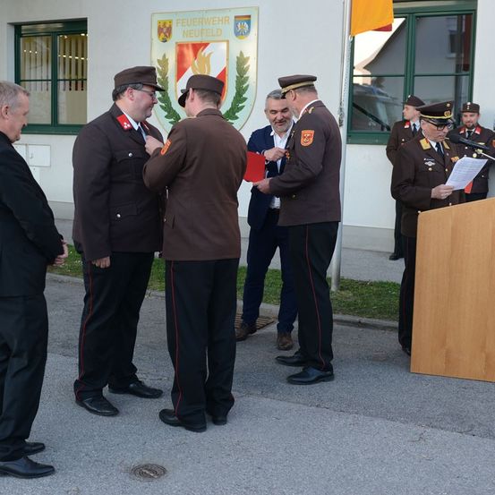 Militärangehörige in braunen Uniformen werden draußen ausgezeichnet. Ein Mann hält eine rote Mappe. Ein Podium steht in der Nähe.