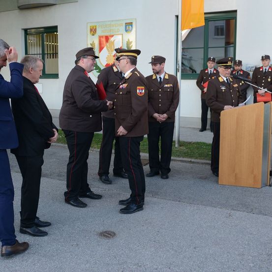 Mehrere Männer in Uniform stehen vor einem Gebäude und schütteln Hände. Ein Mann spricht an einem Podium. Eine Fahne ist im Hintergrund sichtbar.