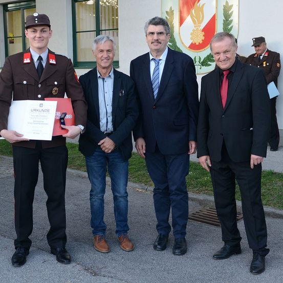 Eine Gruppe von Männern steht vor einem Gebäude. Der Mann in Uniform hält eine Urkunde. Dahinter ist ein Logo mit einer Flamme. Der Mann in der Mitte trägt einen Anzug.