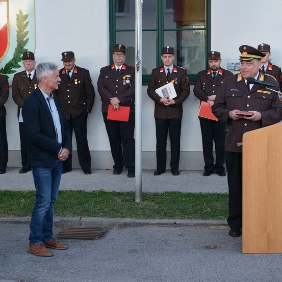 Ein Mann in einem Anzug spricht an einem Podium, während uniformierte Beamte hinter ihm stehen. Die Beamten halten Papiere und Medaillen. Die Veranstaltung findet vor einem Gebäude statt.