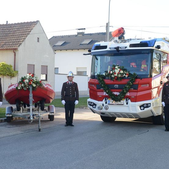 Ein Feuerwehrauto steht auf der Straße, mit Blumen darauf und ein Feuerwehrmann steht neben einem Boot auf einem Anhänger. Dahinter stehen Häuser und eine Straßenlampe.