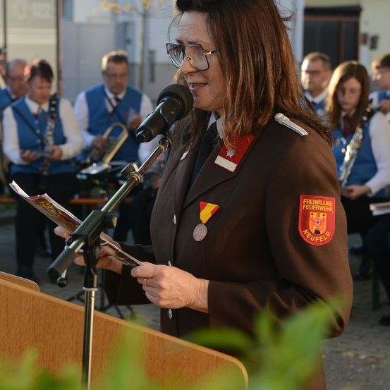 Eine Frau mit Brille und Uniform mit Abzeichen steht an einem Rednerpult und spricht in ein Mikrofon. Hinter ihr spielt eine Band Musikinstrumente.