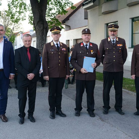 Fünf Männer stehen in Uniform, einer hält eine blaue Mappe, vor einem Gebäude. Dahinter ist ein Baum und eine Menge zu sehen.