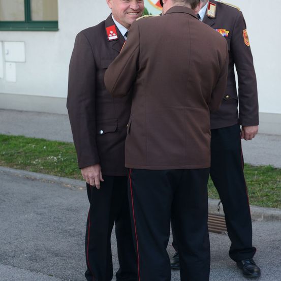 Drei Männer in braunen Uniformen stehen auf einer Straße, einer scheint mit einem anderen zu sprechen. Sie stehen vor einem Gebäude mit einem Logo an der Wand.
