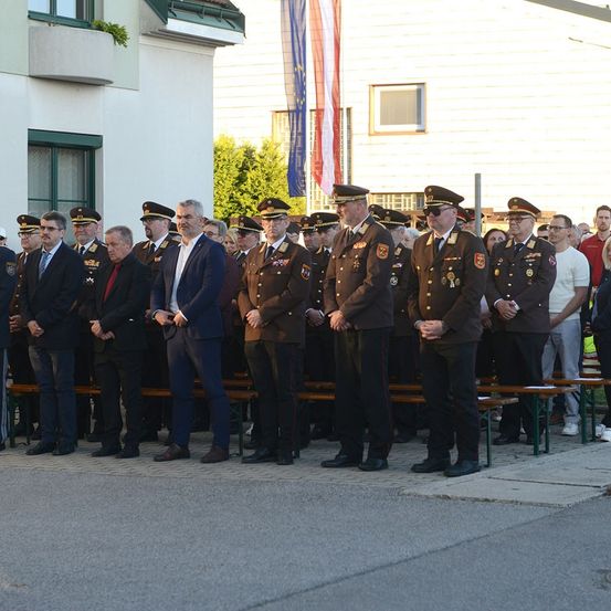 Eine Gruppe von Menschen, einschließlich uniformierter Beamter, steht in einer Reihe. Einige tragen Hüte und Brillen. Das Gebäude hinter ihnen hat eine Flagge und ein Fenster. Bäume sind im Hintergrund.