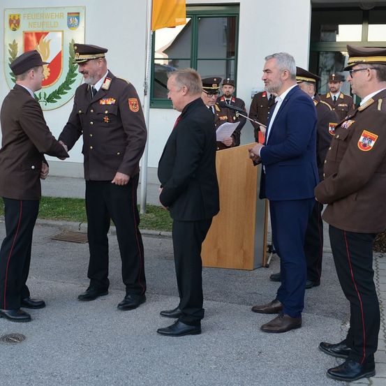 Eine Gruppe von Männern in Uniformen und Anzügen versammelt sich vor einem Gebäude. Ein Mann in einem Anzug spricht an einem Podium.