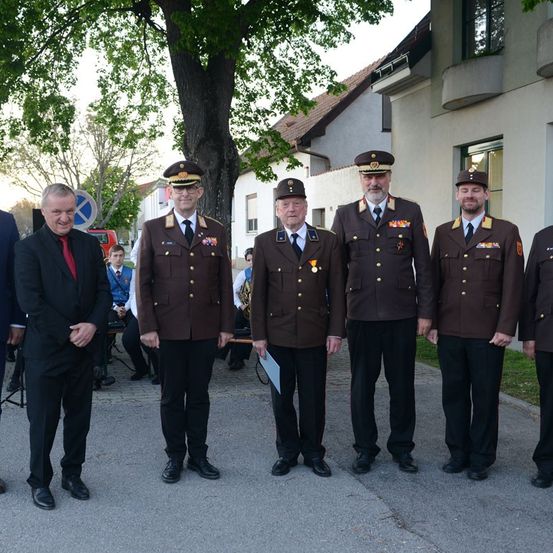 Fünf Männer in Uniform, wahrscheinlich Militäroffiziere, stehen vor einem Gebäude. Sie posieren für ein Foto mit einem Mann in Anzug und Krawatte. Dahinter sind Bäume und ein Gebäude.