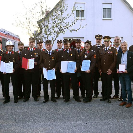 Eine Gruppe uniformierter Personen, darunter Feuerwehrleute und Beamte, posiert für ein Foto vor einem Gebäude. Sie halten Urkunden und Auszeichnungen in den Händen, und einige tragen Mützen und Brillen. Ein Feuerwehrwagen steht hinter ihnen.