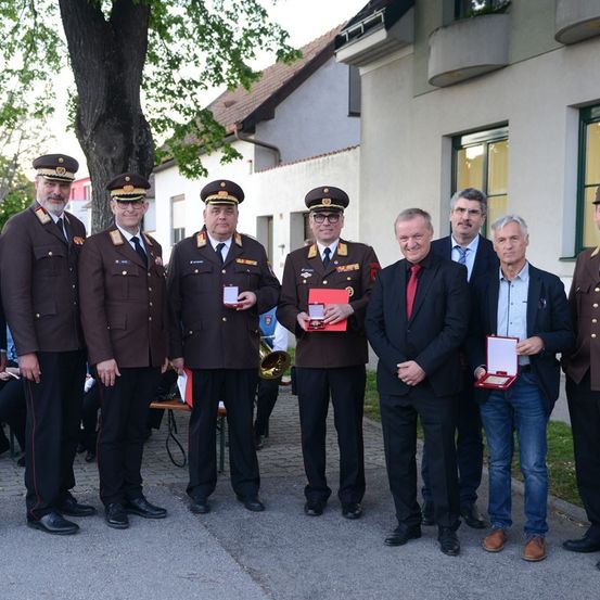 Mehrere Männer in Uniformen und Anzügen stehen in einer Reihe und halten Auszeichnungen. Ein Mann spielt Trompete. Ein Baum und Gebäude sind im Hintergrund.