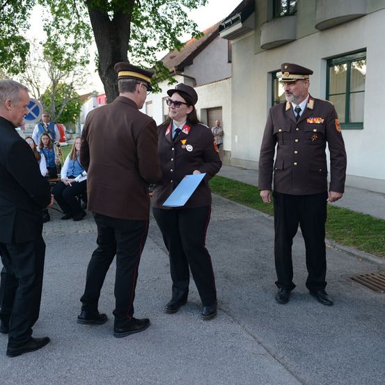 Eine Frau in Uniform hält ein Dokument und steht zwischen zwei Männern in Uniform, während mehrere Menschen dahinter sitzen, einer spielt Posaune.