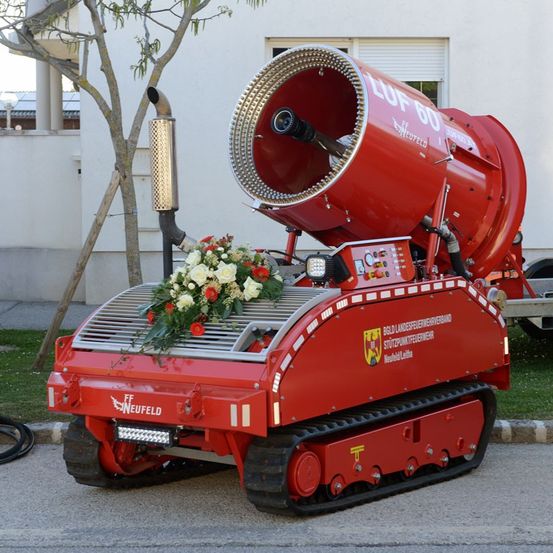 Ein rotes Fahrzeug mit Blumen und der Aufschrift 'LUF 60' und 'Neufeld' steht am Straßenrand. Es hat einen großen Lüfter vorne.