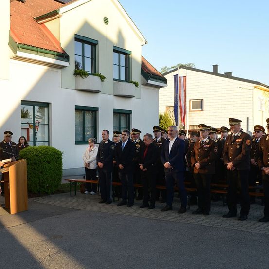 Ein formeller Anlass mit uniformierten Beamten und Würdenträgern vor einem Gebäude mit Flaggenmast und Bänken.