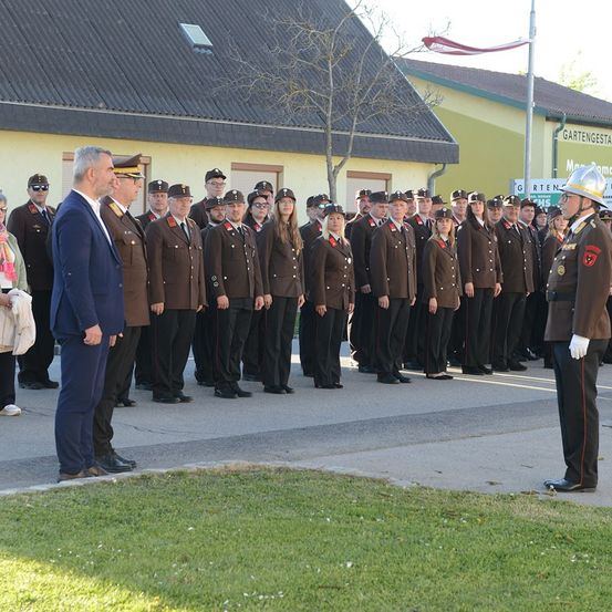 Eine Gruppe uniformierter Personen steht in einer Reihe, mit einer Flagge und einem Gebäude im Hintergrund. Ein Mann im Anzug und ein Militäroffizier sind vorne.