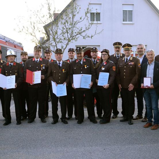 Eine Gruppe uniformierter Beamter, einschließlich Feuerwehrleute und Militärangehöriger, steht vor einem weißen Haus. Sie halten Urkunden und Medaillen.