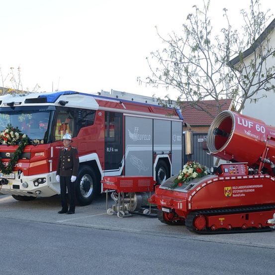 Ein Feuerwehrauto steht vor einem Gebäude mit einem Mann in Uniform neben ihm. Das Auto ist mit Kränzen und Blumen geschmückt, und ein roter und weißer Feuerwehrwagen steht dahinter.