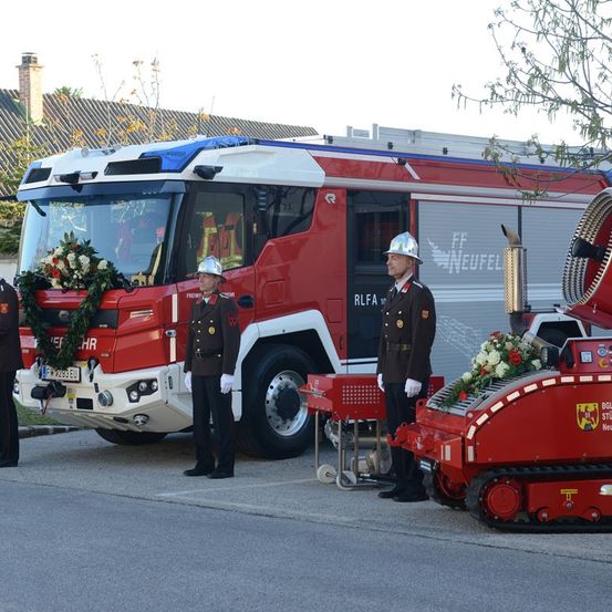 Zwei Feuerwehrleute stehen neben einem roten Feuerwehrauto mit einer Girlande, ein weiterer steht bei einem roten gepanzerten Fahrzeug mit Blumen.