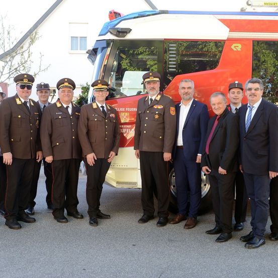 Eine Gruppe uniformierter Männer steht vor einem roten Feuerwehrauto. Sie alle lächeln und scheinen für ein Foto zu posieren.