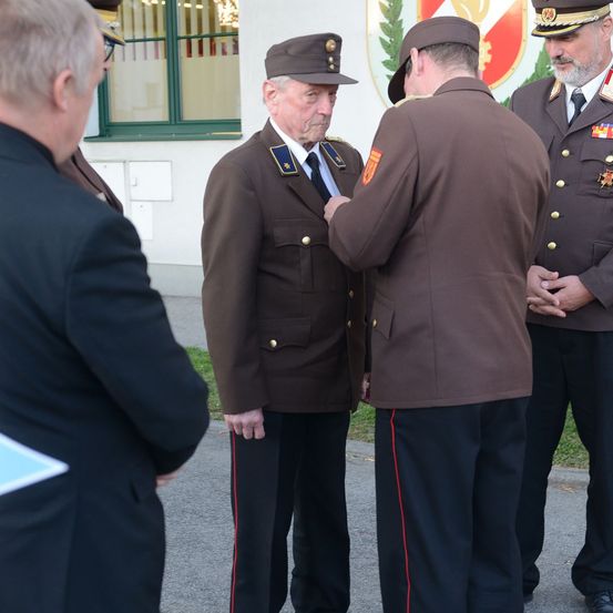 Ein älterer Mann in Uniform erhält eine Medaille von einem jüngeren Mann in ähnlicher Uniform. Sie stehen vor einem Gebäude.