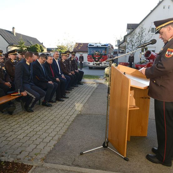 Ein Mann in Uniform spricht in ein Mikrofon an einem Rednerpult, während andere hinter ihm sitzen. Ein Feuerwehrwagen steht im Hintergrund auf der Straße.
