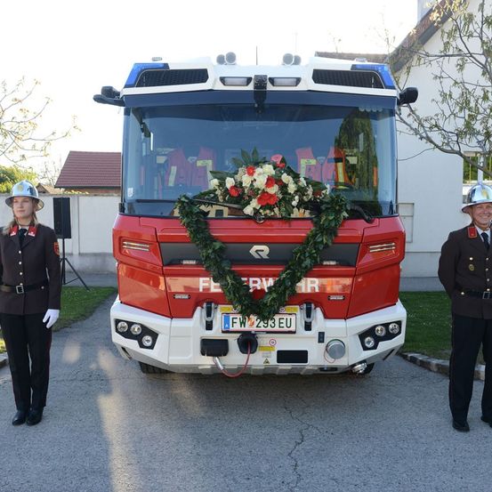 Zwei Feuerwehrleute in Uniform stehen vor einem roten Feuerwehrauto mit Blumen auf der Vorderseite.