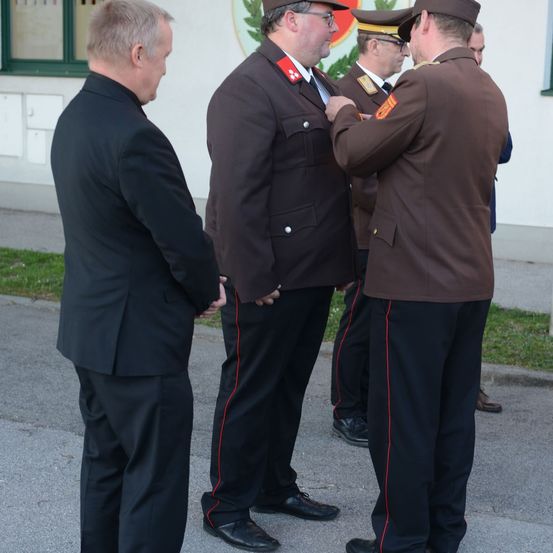 Ein formelles Ereignis mit zwei uniformierten Männern und zwei Männern in Anzügen; ein uniformierter Mann befestigt eine Medaille an einem anderen.